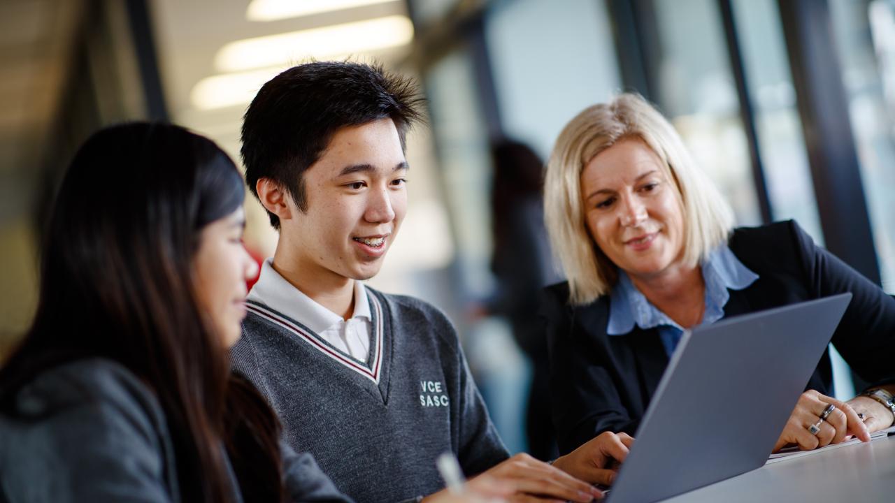 Teacher sitting with two high school students looking at a laptop. 