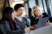 Teacher sitting with two high school students looking at a laptop. 